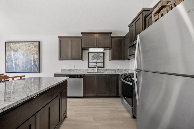 kitchen with light stone counters, dark brown cabinetry, a sink, appliances with stainless steel finishes, and light wood-type flooring