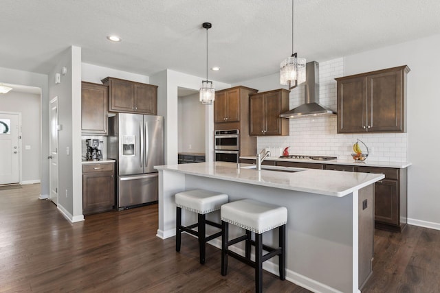 kitchen featuring a center island with sink, stainless steel appliances, hanging light fixtures, a sink, and wall chimney exhaust hood