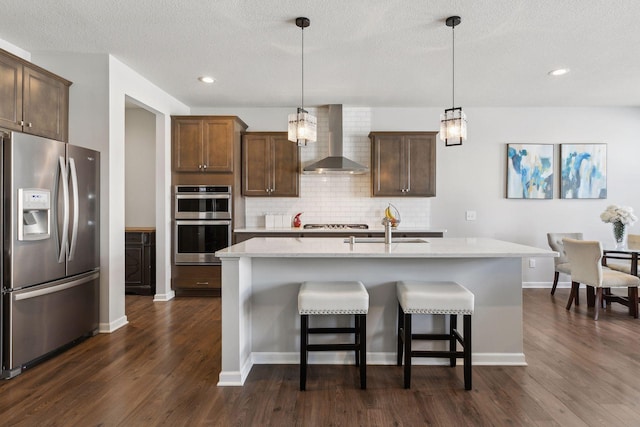 kitchen with appliances with stainless steel finishes, a kitchen island with sink, wall chimney range hood, and decorative light fixtures