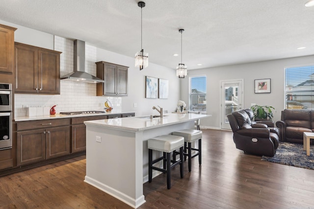 kitchen featuring a center island with sink, wall chimney exhaust hood, dark wood-type flooring, a sink, and a kitchen bar