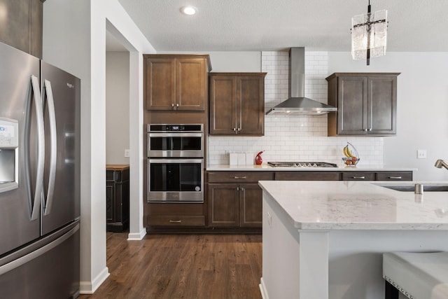 kitchen with dark wood-style flooring, light stone countertops, stainless steel appliances, wall chimney range hood, and pendant lighting