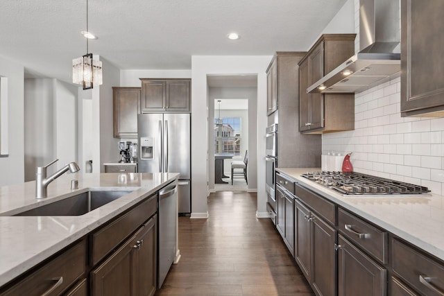 kitchen with light stone counters, pendant lighting, stainless steel appliances, a sink, and wall chimney exhaust hood