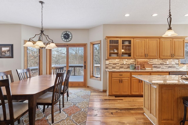 kitchen with light stone counters, hanging light fixtures, glass insert cabinets, light wood-style floors, and backsplash