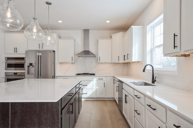 kitchen with hanging light fixtures, stainless steel appliances, white cabinets, wall chimney range hood, and sink