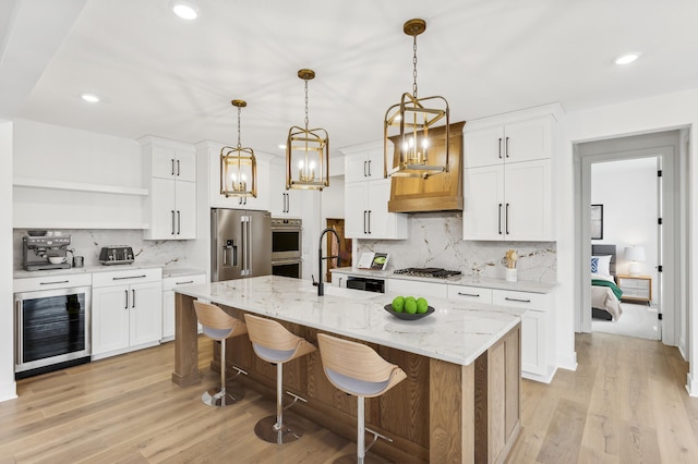 kitchen featuring a kitchen island with sink, stainless steel appliances, decorative light fixtures, white cabinets, and beverage cooler