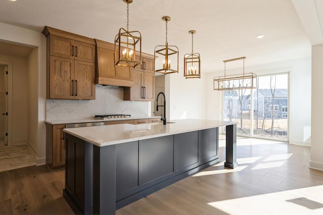 kitchen with brown cabinets, a center island with sink, tasteful backsplash, wood finished floors, and stainless steel gas stovetop