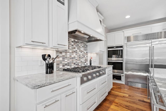 kitchen featuring stainless steel appliances, wood finished floors, white cabinetry, backsplash, and custom range hood