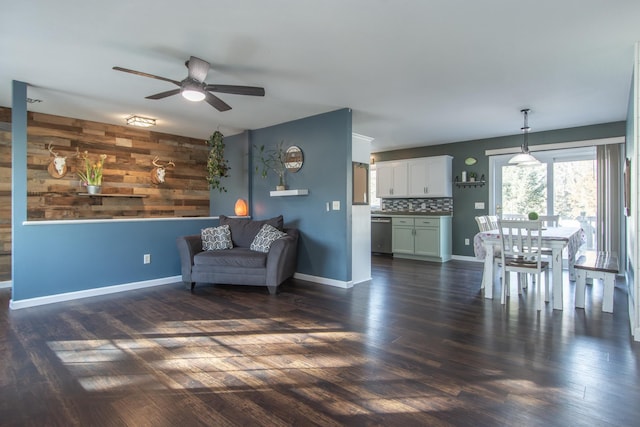 living area featuring dark wood-type flooring, wooden walls, baseboards, and ceiling fan