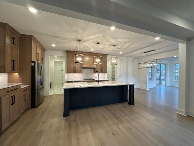 kitchen with an island with sink, decorative backsplash, light wood-style flooring, brown cabinetry, and stainless steel fridge