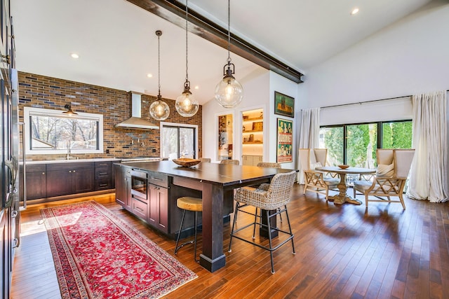 kitchen with high vaulted ceiling, beam ceiling, hanging light fixtures, dark wood-type flooring, and wall chimney range hood