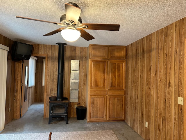 interior space featuring ceiling fan, a wood stove, a textured ceiling, and wooden walls