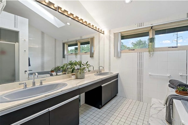 full bath featuring double vanity, vaulted ceiling with skylight, tile walls, and a sink