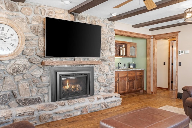 living area with beam ceiling, wood finished floors, a stone fireplace, baseboards, and ceiling fan