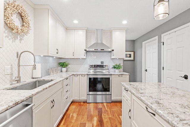 kitchen with wall chimney range hood, appliances with stainless steel finishes, a sink, white cabinets, and light wood-style floors
