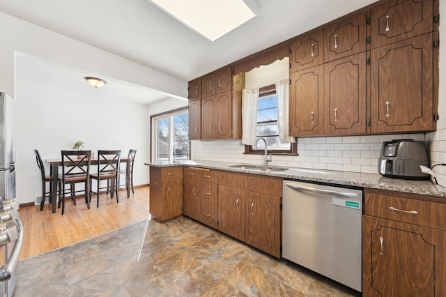 kitchen with stainless steel appliances, a sink, backsplash, light stone countertops, and brown cabinetry