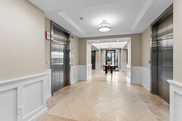 corridor with a wainscoted wall, a tray ceiling, light tile patterned flooring, and elevator