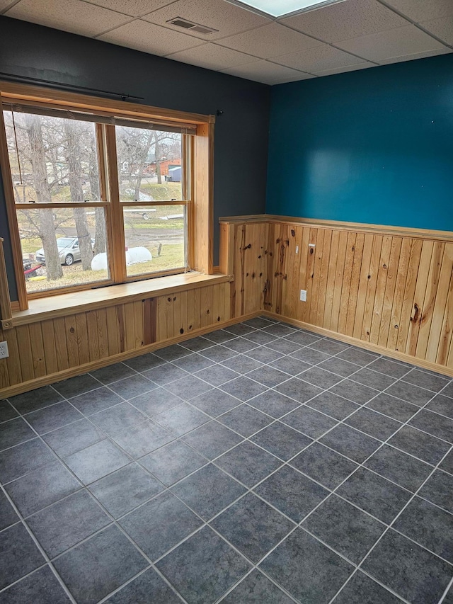 unfurnished room with visible vents, wainscoting, wooden walls, a drop ceiling, and baseboards