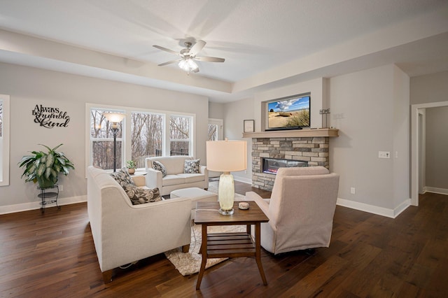 living area with a stone fireplace, dark wood-style flooring, a ceiling fan, and baseboards