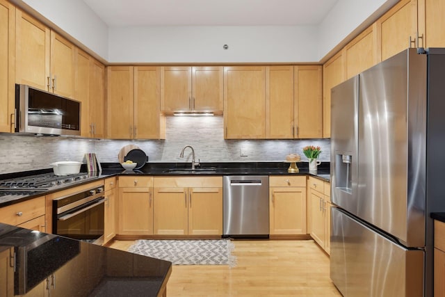 kitchen with light wood finished floors, stainless steel appliances, dark countertops, light brown cabinetry, and a sink