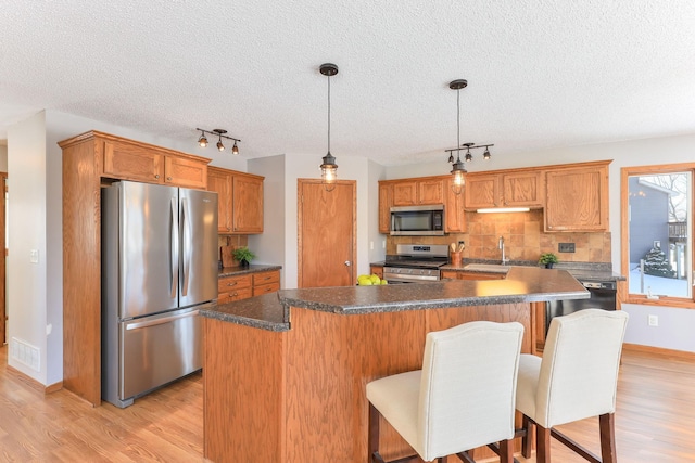 kitchen with stainless steel appliances, brown cabinetry, dark countertops, and decorative light fixtures