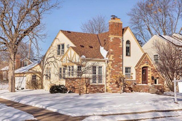 tudor-style house featuring brick siding, a shingled roof, a chimney, and stucco siding