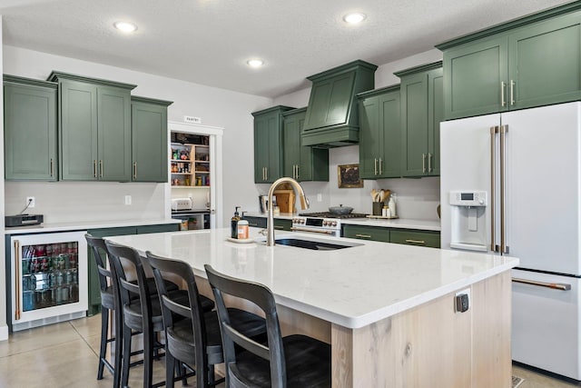 kitchen with light stone counters, wine cooler, a center island with sink, high end white fridge, and green cabinets