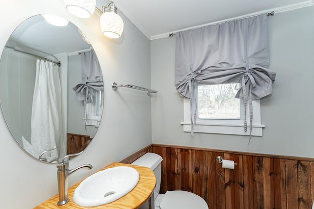 bathroom featuring wooden walls, toilet, a wainscoted wall, ornamental molding, and a sink