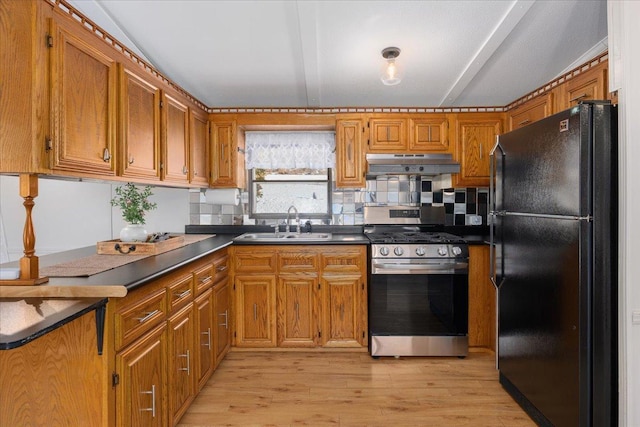 kitchen with under cabinet range hood, a sink, stainless steel range with gas cooktop, freestanding refrigerator, and brown cabinetry