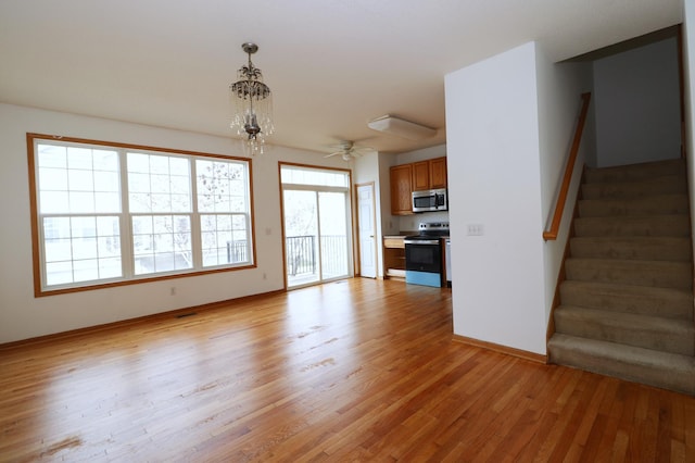 unfurnished living room with baseboards, stairway, light wood finished floors, and ceiling fan with notable chandelier