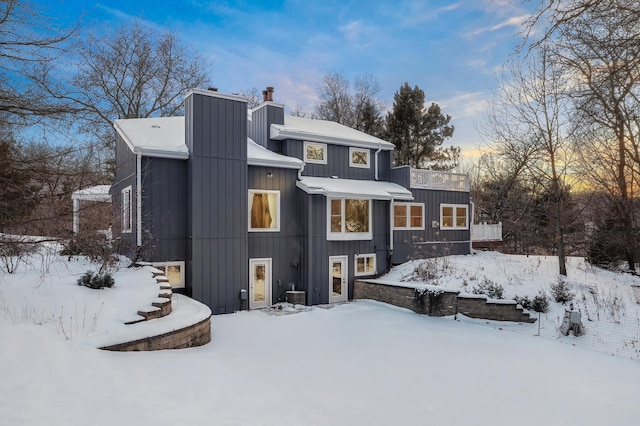 snow covered house with stairs, a chimney, and board and batten siding