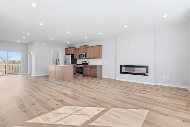 kitchen with a kitchen island with sink, a sink, open floor plan, appliances with stainless steel finishes, and a glass covered fireplace