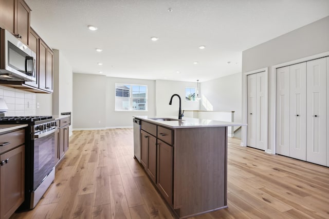 kitchen featuring light wood finished floors, decorative backsplash, a kitchen island with sink, stainless steel appliances, and a sink