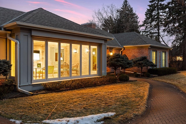 back of property at dusk featuring brick siding, roof with shingles, a chimney, and a sunroom