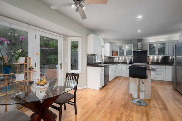 kitchen with dark countertops, glass insert cabinets, wall chimney range hood, light wood-style flooring, and appliances with stainless steel finishes