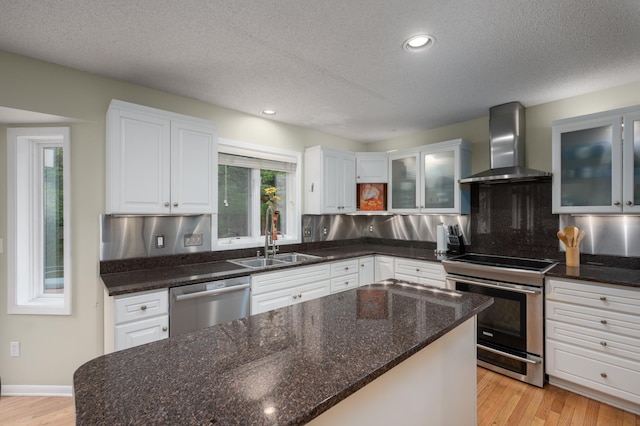 kitchen featuring wall chimney range hood, appliances with stainless steel finishes, light wood-style floors, white cabinetry, and a sink