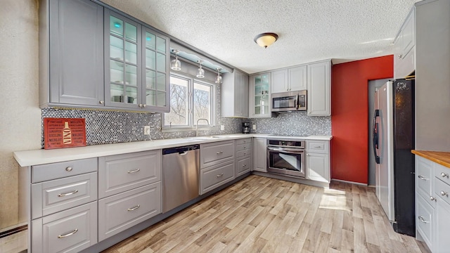 kitchen with light wood-style flooring, stainless steel appliances, a sink, tasteful backsplash, and glass insert cabinets