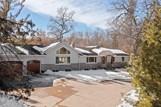 snow covered rear of property featuring a chimney, stone siding, an attached garage, and concrete driveway