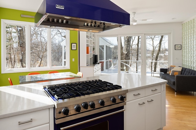 kitchen featuring high end stove, a ceiling fan, ventilation hood, light wood-style floors, and white cabinets