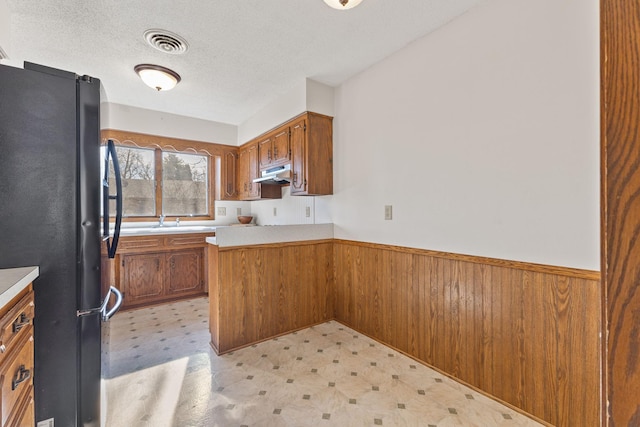 kitchen featuring a wainscoted wall, under cabinet range hood, freestanding refrigerator, brown cabinetry, and light floors