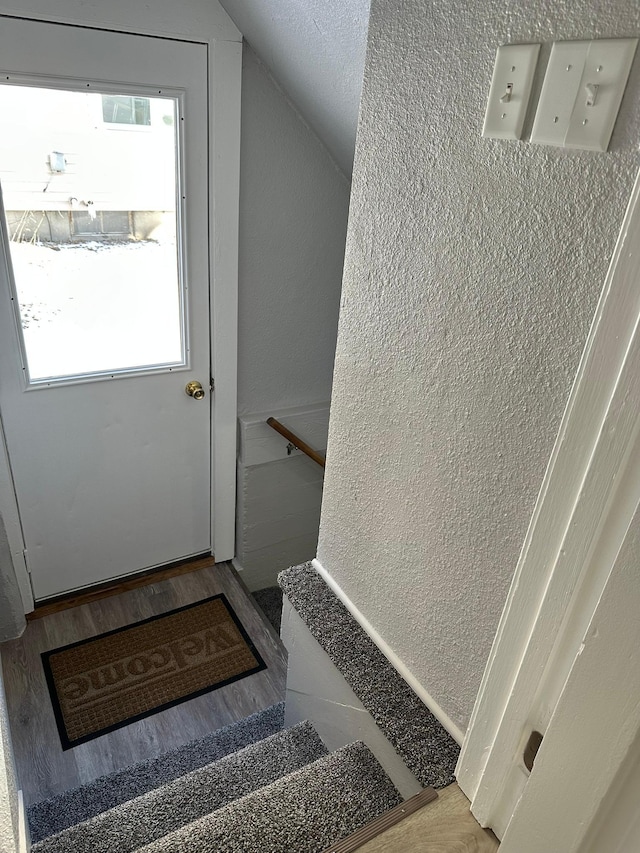 stairs with a wealth of natural light, vaulted ceiling, wood finished floors, and a textured wall