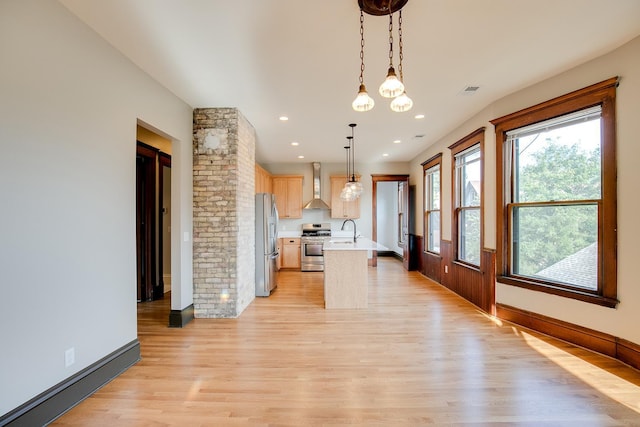 kitchen with decorative light fixtures, light countertops, appliances with stainless steel finishes, wall chimney exhaust hood, and a center island with sink