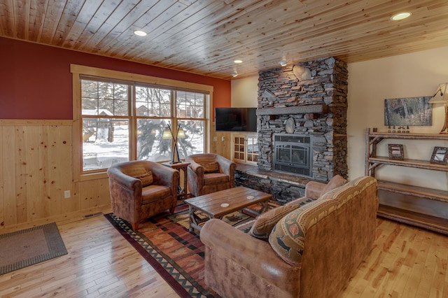 living area with wooden walls, wainscoting, wood ceiling, a stone fireplace, and light wood-style floors