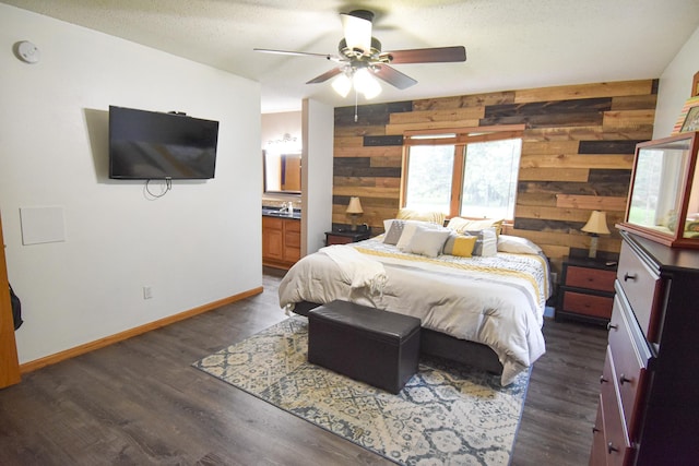 bedroom with a textured ceiling, dark wood-type flooring, wood walls, baseboards, and ensuite bath