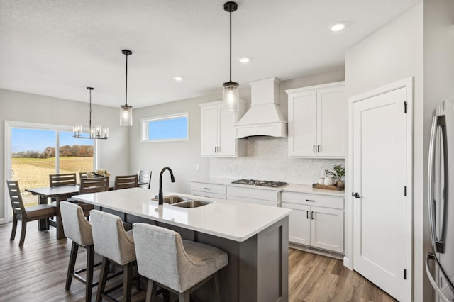 kitchen featuring white cabinets, custom range hood, a kitchen island with sink, pendant lighting, and a sink