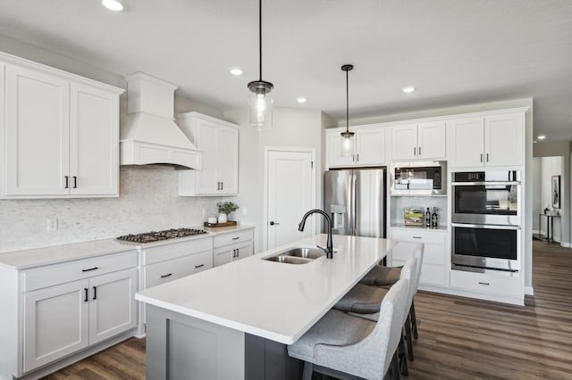 kitchen featuring premium range hood, light countertops, appliances with stainless steel finishes, an island with sink, and decorative light fixtures