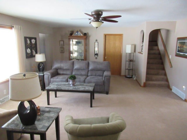 living area with ceiling fan, stairway, baseboards, and light colored carpet