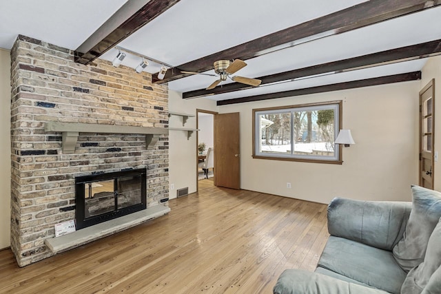 living room featuring visible vents, light wood-style flooring, a ceiling fan, a brick fireplace, and beamed ceiling