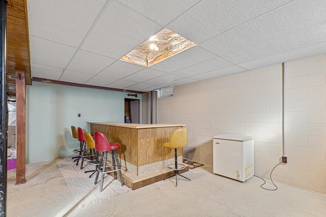 bar featuring concrete block wall, a dry bar, a drop ceiling, unfinished concrete flooring, and fridge