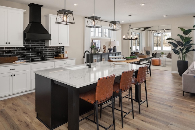 kitchen featuring decorative light fixtures, premium range hood, a sink, black stovetop, and a center island with sink