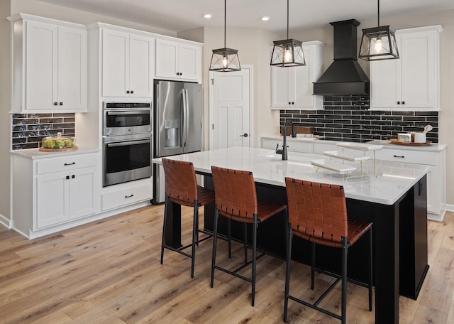 kitchen featuring pendant lighting, appliances with stainless steel finishes, a kitchen island with sink, white cabinets, and premium range hood